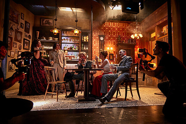 Eine Gruppe von fünf Personen sitzt und steht in einem stilvoll eingerichteten Raum mit zwei Kameraleuten, die die Szene filmen. Der Raum ist dekoriert mit vintage Möbeln und Tapeten, was an eine Bar oder ein Café in einer vergangenen Ära erinnert. Zwei Personen sitzen an einem Tisch, während die anderen drei stehen, alle in eleganter Kleidung. Die Beleuchtung ist warm, was dem Bild eine dramatische und nostalgische Stimmung verleiht. Im Hintergrund sind Regale mit verschiedenen Gegenständen und Getränken z