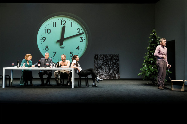 Szene aus einer Theateraufführung mit fünf Personen am Tisch vor einer großen Uhr.
