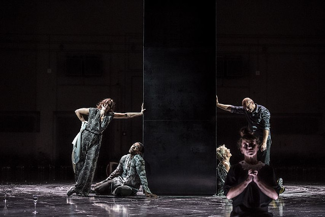 Eine Gruppe von fünf Personen posiert in dramatischer Beleuchtung um eine monumentale, schwarze Säule. Zwei von ihnen stützen sich gegen die Säule, während eine Person im Vordergrund mit gefalteten Händen kniet. Die Szene vermittelt eine ernste, theatralische Atmosphäre.