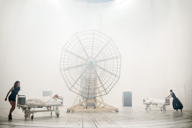 Das Bild zeigt eine futuristische Szene in einem großen, leeren weißen Raum. Im Mittelpunkt steht eine große, ungewöhnlich gestaltete Struktur, die einem riesigen Ventilator ähnelt. Links und rechts davon sind zwei Krankenhausbetten mit weißen Leintüchern. In den Betten liegen Personen, während auf jeder Seite eine Person in schwarzer Kleidung die Betten schiebt. Die gesamte Atmosphäre wirkt surreal und geheimnisvoll.