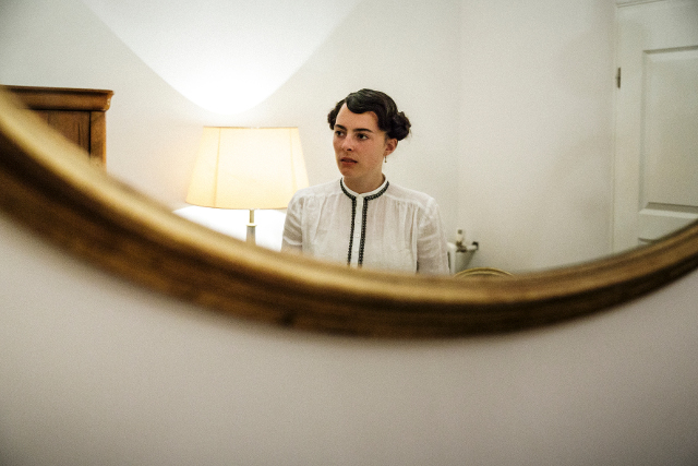 Ein reflektierendes Bild in einem runden Spiegel zeigt eine Person mit kurzem, welligem Haar. Die Person trägt eine weiße Bluse mit einem schwarzen Kragen. Im Hintergrund ist eine Lampe und ein Teil einer weißen Wand zu sehen. Die Stimmung wirkt ruhig und gedämpft durch die dezente Beleuchtung.