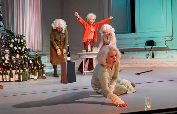 In einem minimalistischen Raum mit einer hellblauen Wand und rosa Vorhängen sind vier ältere Frauen zu sehen. Im Hintergrund steht ein Tisch mit einer Flasche, während eine Frau im orangefarbenen Pullover die Hände triumphierend in die Luft streckt. Neben ihr beugt sich eine andere Frau im beigen Cardigan nach vorne. Ganz vorne im Bild kniet eine Frau in einem hellgrünen Kleid dramatisch auf dem Boden und greift nach einem umgekippten Glas.