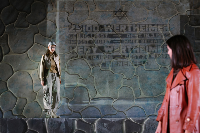 Zwei Personen stehen auf einer Bühne mit einer Wand, die wie Stein aussieht. Eine Person trägt einen Hut und steht auf einer hohen Plattform. Die andere Person trägt einen langen Mantel und sieht zur ersten Person hinüber. Im Hintergrund ist ein verblassener Text mit einem Davidstern sichtbar.