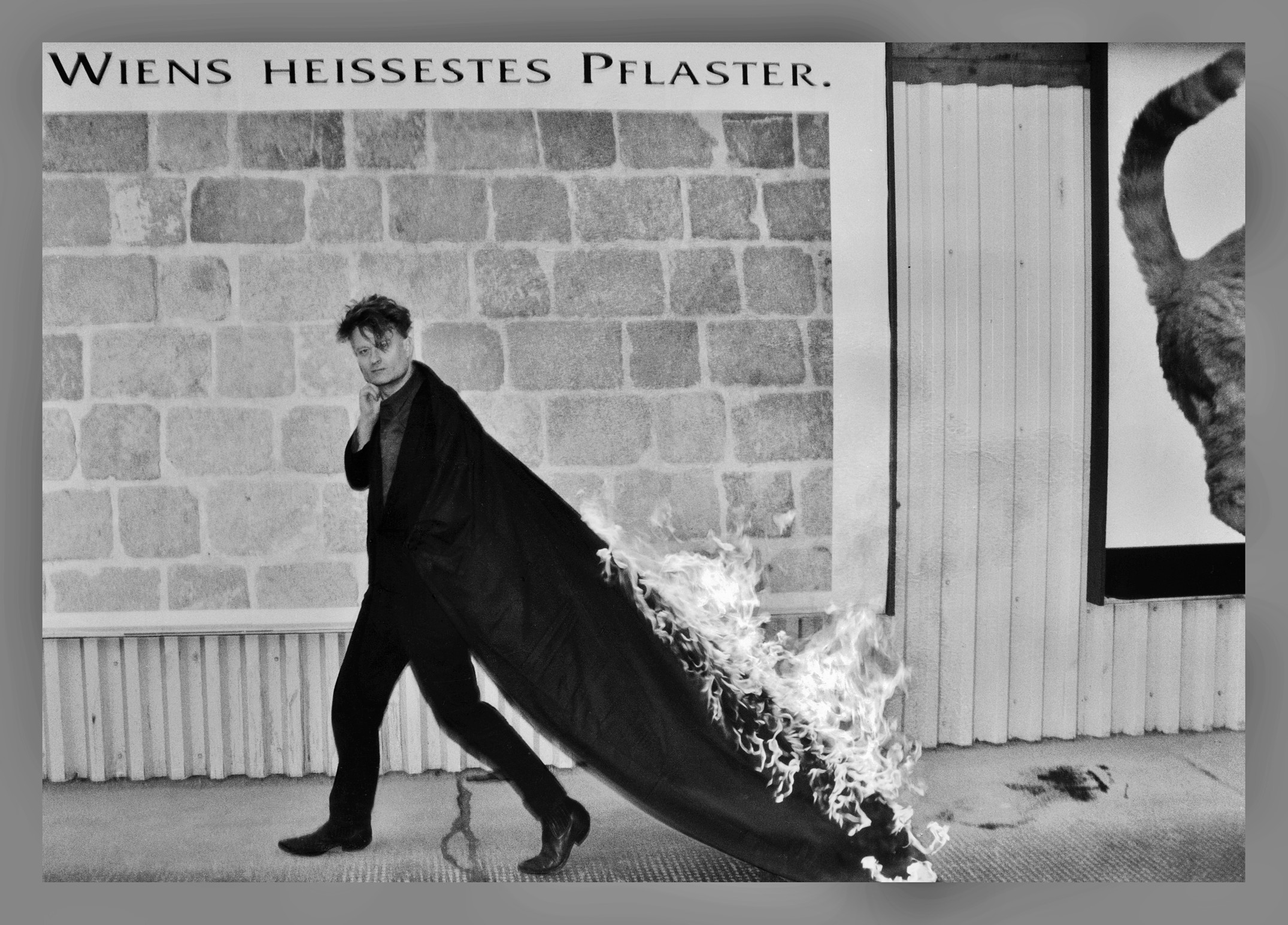 Ein eleganter Mann in einem schwarzen Anzug geht selbstbewusst, während sein langer Mantel hinter ihm lodert. Hinter ihm ist eine Mauer mit der Aufschrift 
