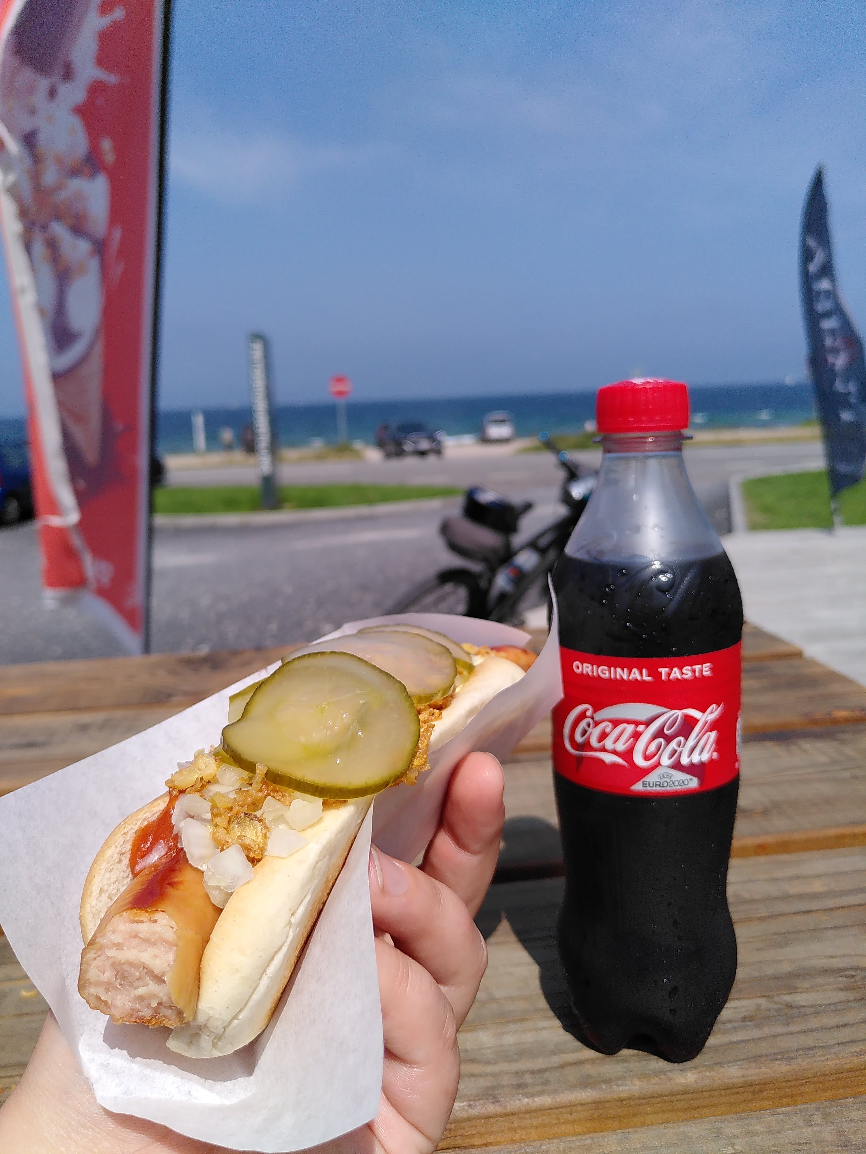 Foto von einem Picknicktisch am Strand. Eine Hand hält einen Hotdog, daneben steht eine kleine Cola-Flasche.