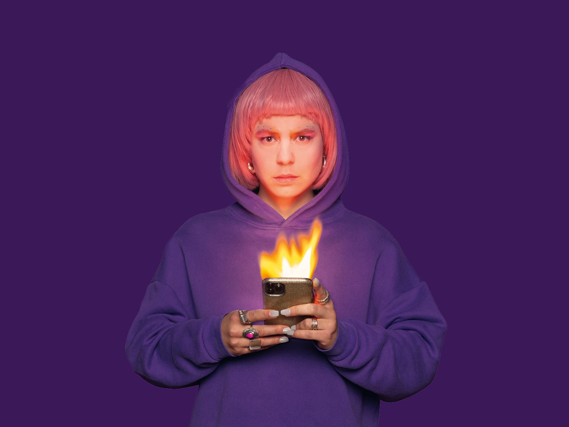 Produktionsfoto:eine junge Frau mit pinken Haaren und einem lila Kapuzenpullover schaut frontal in die Kamera und  hält ein brennendes Smartphone in der Hand