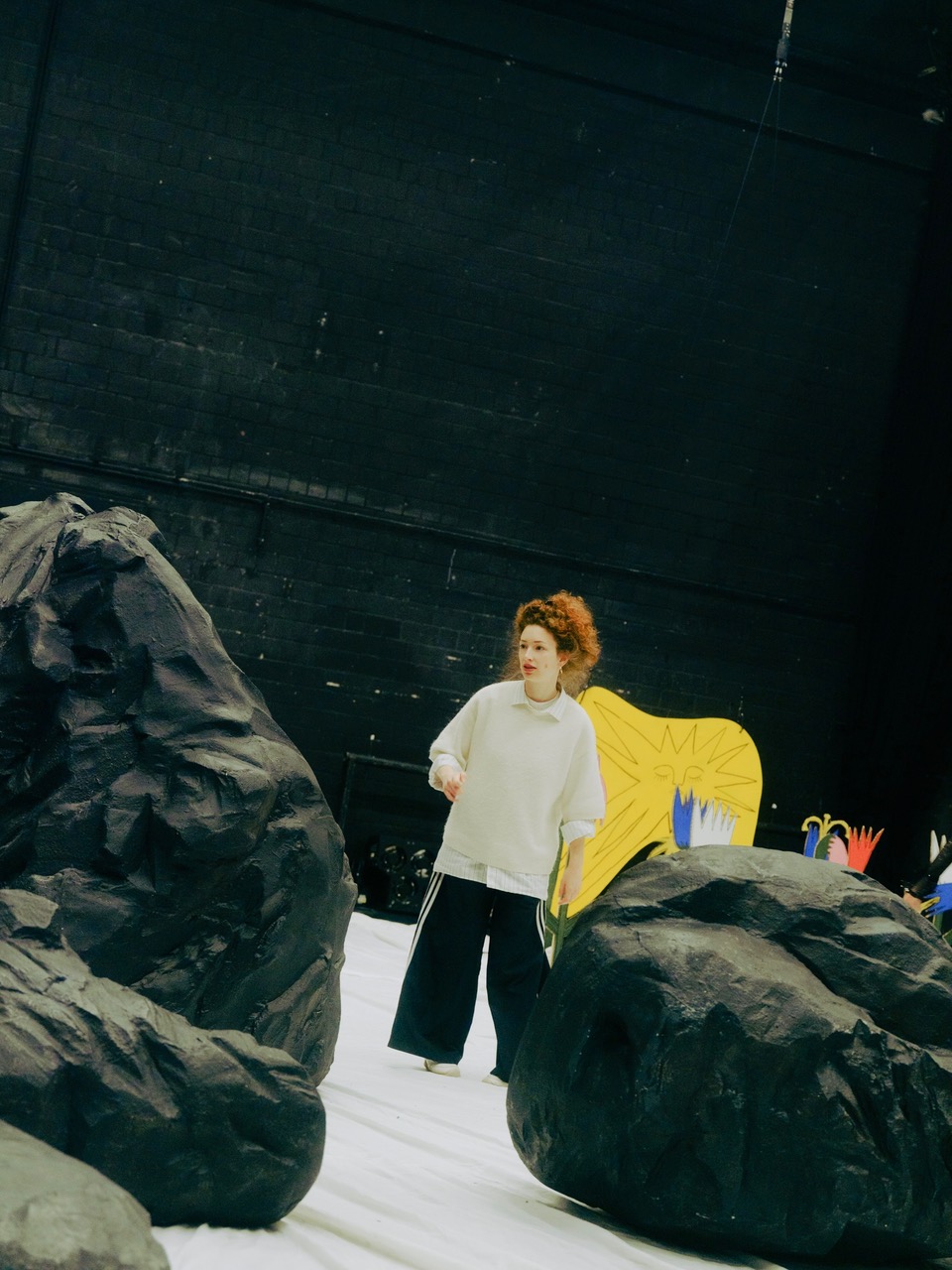 Regisseurin Lea Marlen Balzer inmitten des Bühnenbildes bestehend aus schwarzen Felsformationen. Sie trägt einen weißen Strickpulli und eine weitgeschnittene schwarze Hose