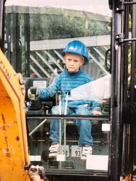 Leo Meier als Kind auf einem gelben Bagger. Er trägt einen hellblauen Helm und schaut sehr konzentriert auf die Baggerschaufel vor ihm. 