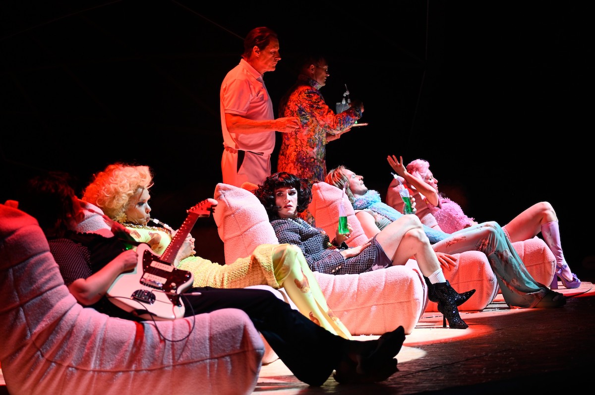 Das Ensemble fläzt sich in eine pinke Sofalandschaft, während ein Mann im Seidenmantel im Halbschatten Getränke auf einem Tablett bugsiert.