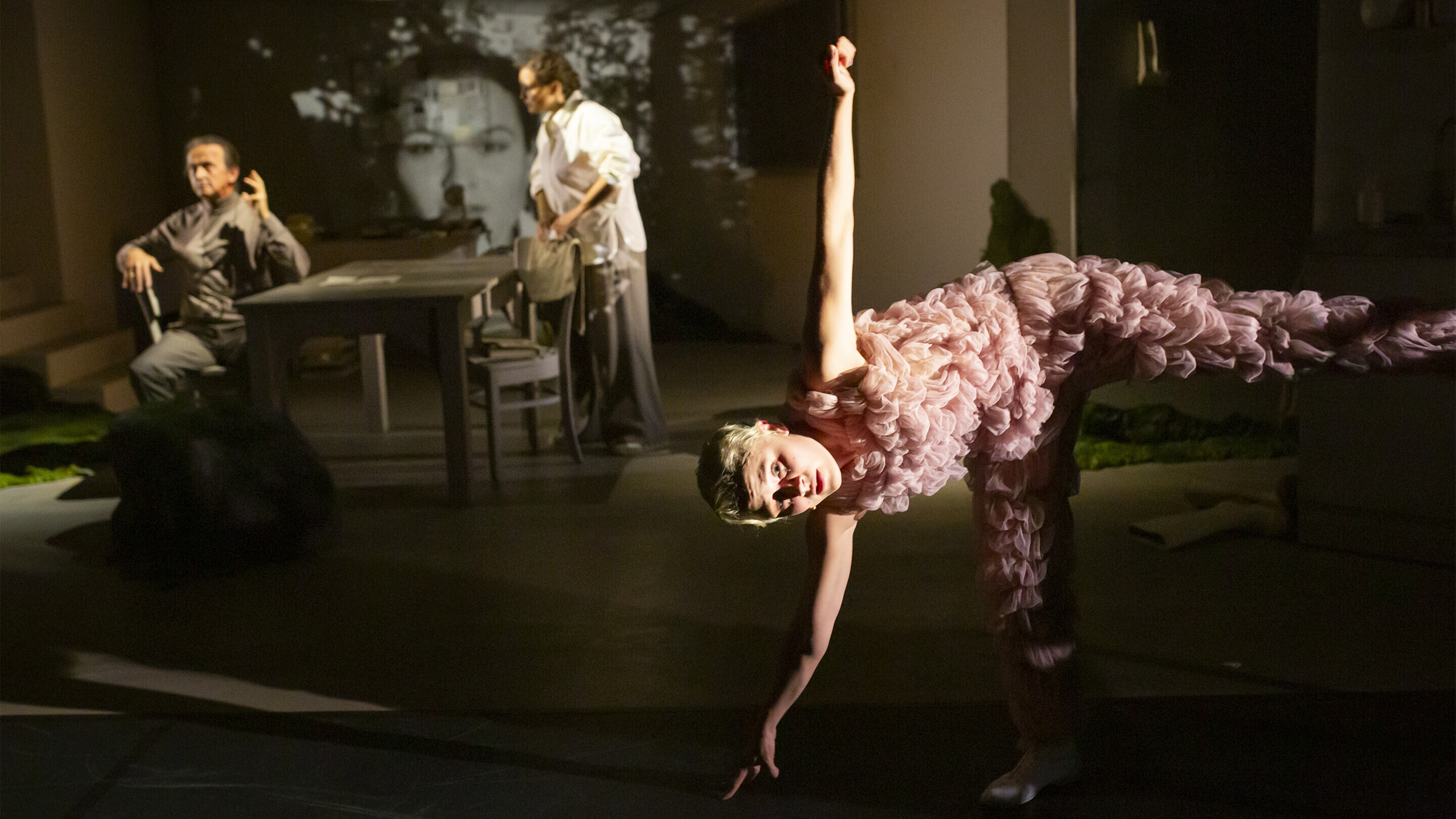 Eine junge Frau mit kurzen, blonden Haaren balanciert auf einem Bein und berührt mit ihrer rechten Hand den Bühnenboden. Im Hintergrund ein Mann und eine Frau mittleren Alters am Tisch sitzend. Sie sind mit ihrer eigenen Szene beschäftigt und ignorieren die Frau im Bildvordergrund. 