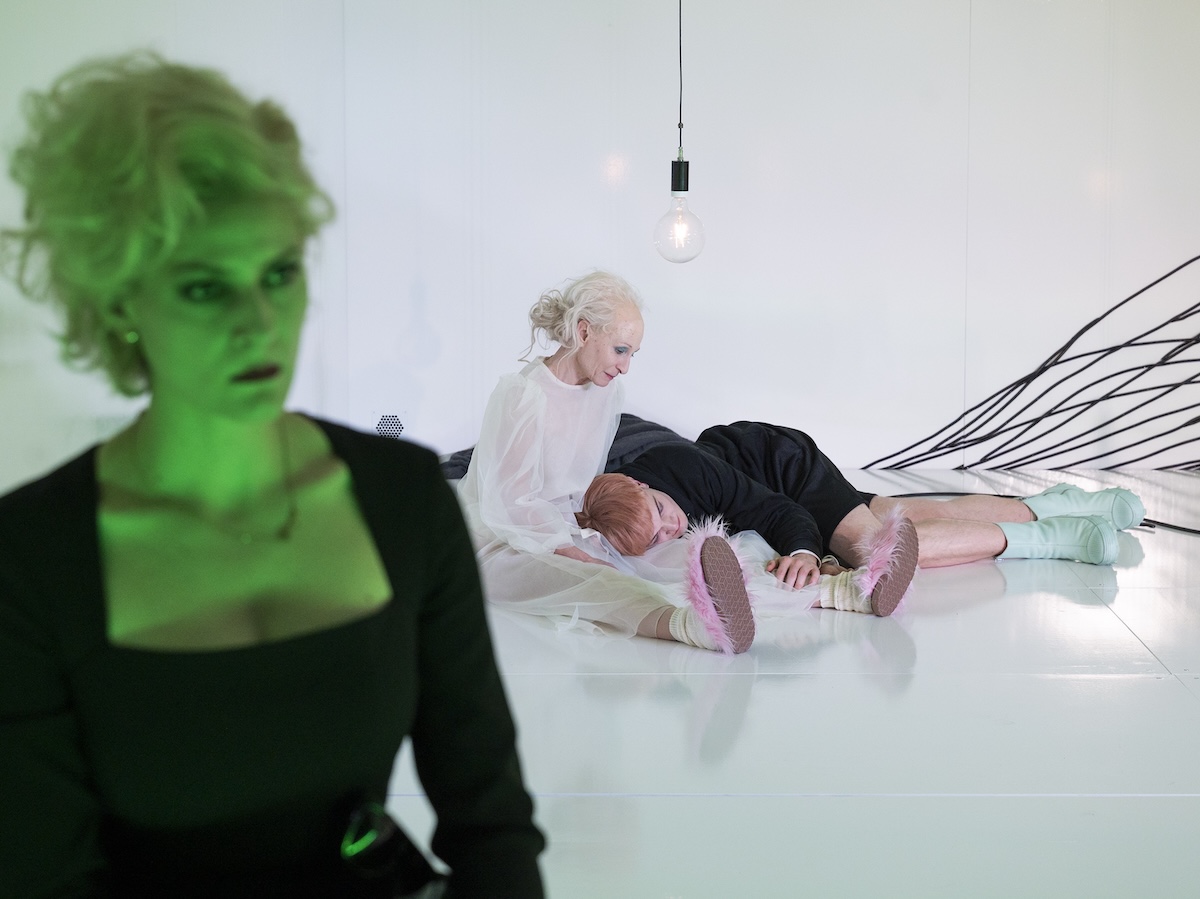 Drei Personen in einem Theaterstück auf einer weißen Bühne. Eine blonde Frau im Vordergrund blickt aus dem Bild. Dahinter liegen zwei Personen, die eine hält die andere.