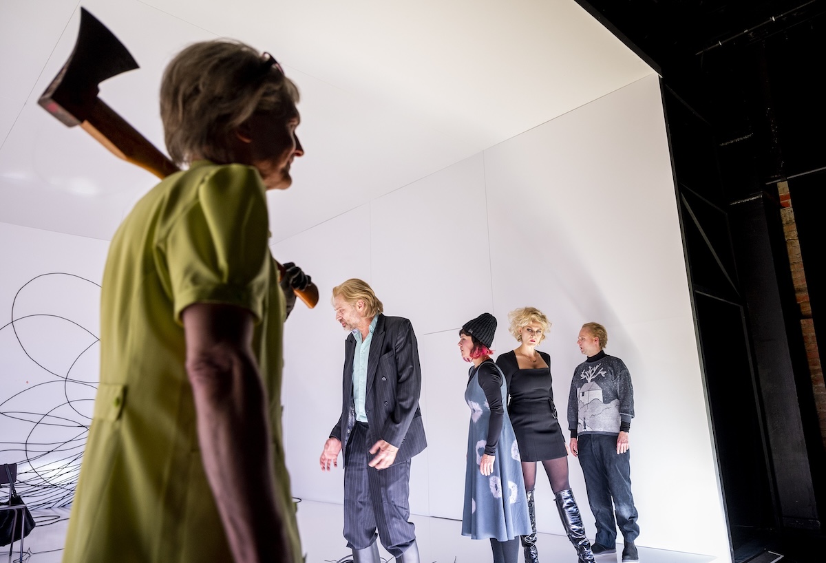 Ein Theaterfoto: Eine ältere Frau mit Axt in gelbgrünem Kleid steht links im Vordergrund, schaut nach rechts. Rechts stehen vier Personen (Mann, Frau, Frau, Mann) auf einer weißen Bühne.