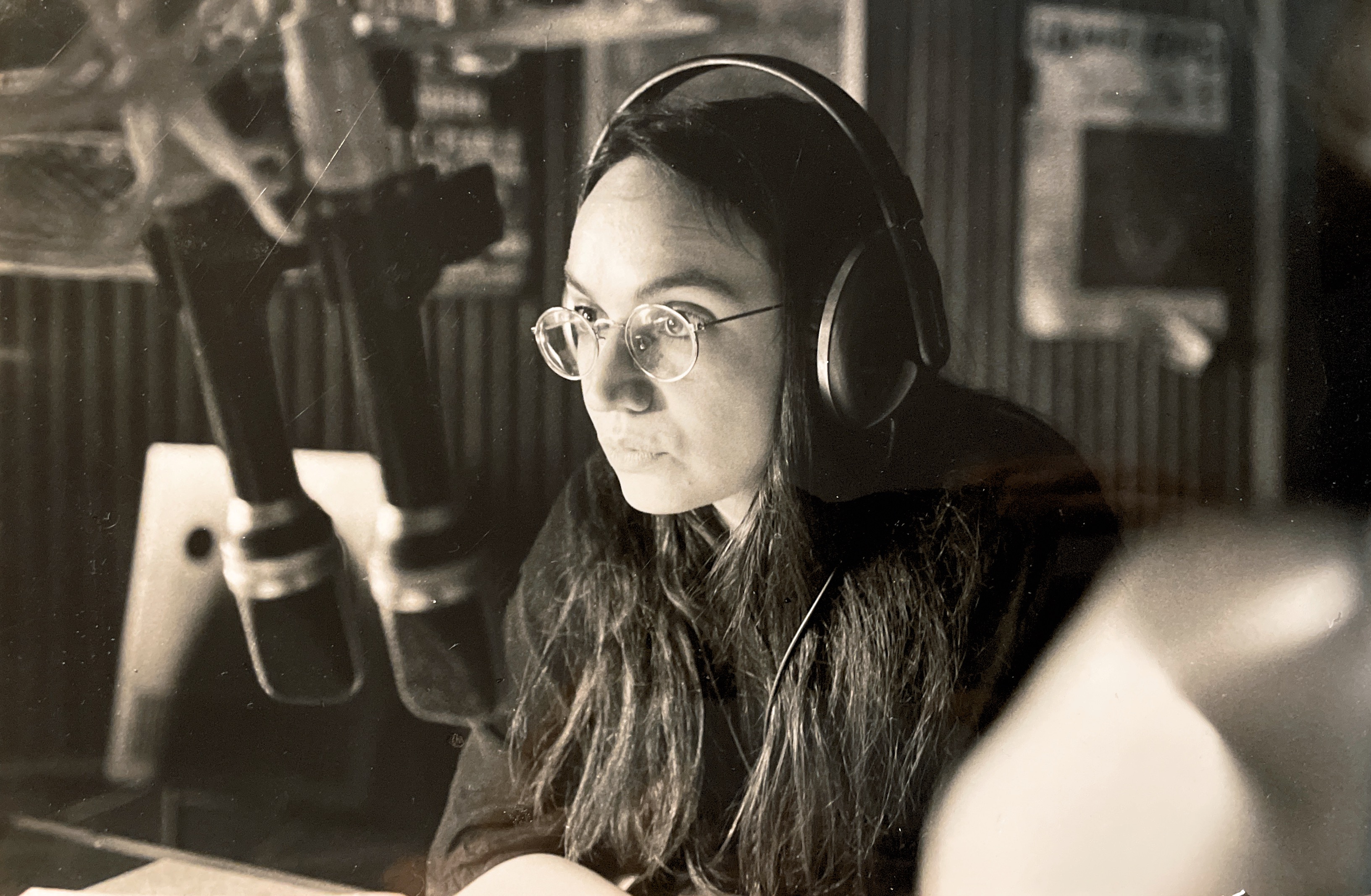 Eine junge Frau mit Brille und langen, dunklen Haaren sitzt mit Kopfhörern im Profil an einem Mikrofon in einem schwarz-weiß Foto.