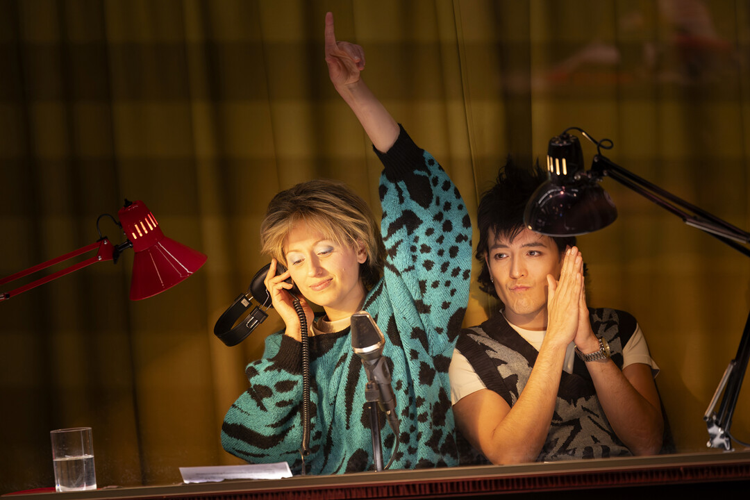 Zwei junge Erwachsene sitzen in einem Tonstudio: Eine Frau telefoniert mit hochgerecktem Finger, ein Mann blickt mit den Händen vor der Brust zu ihr, beide unter Schreibtischlampen.