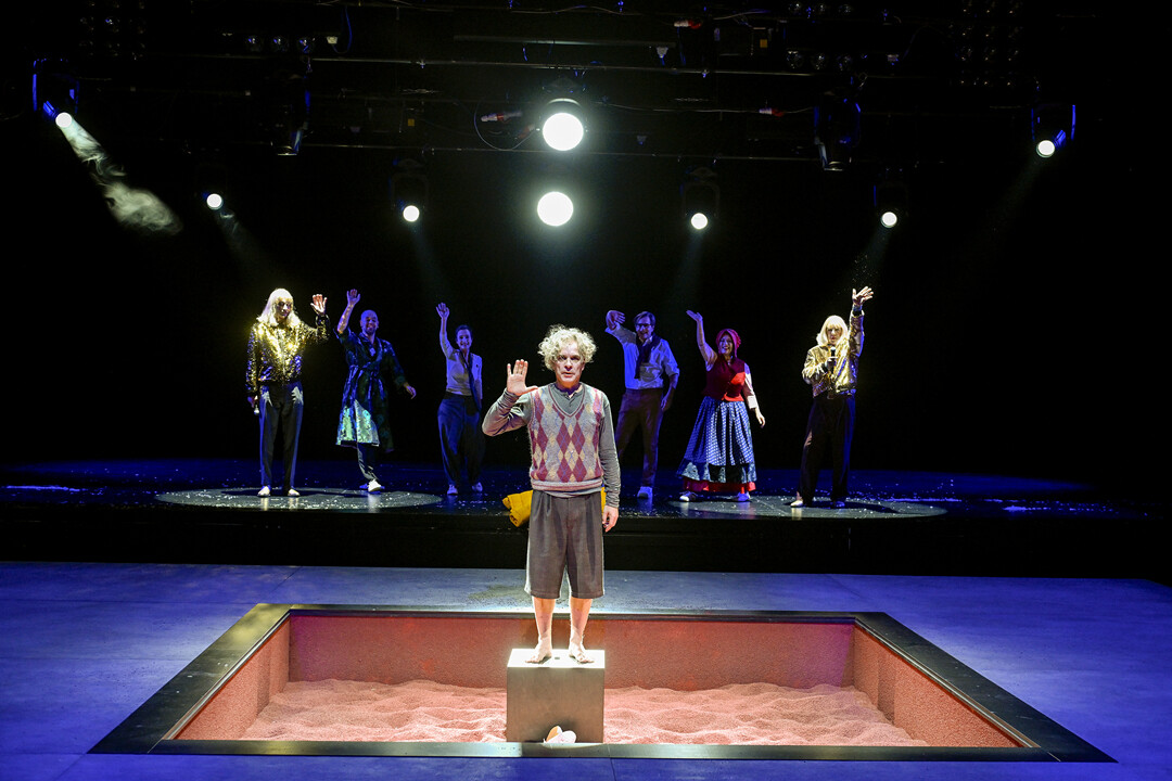 Junge in der Mitte einer Bühne, umgeben von fünf Darstellenden mit Kostümen und winkenden Händen im Rampenlicht, vor einer schwarzen Kulisse.