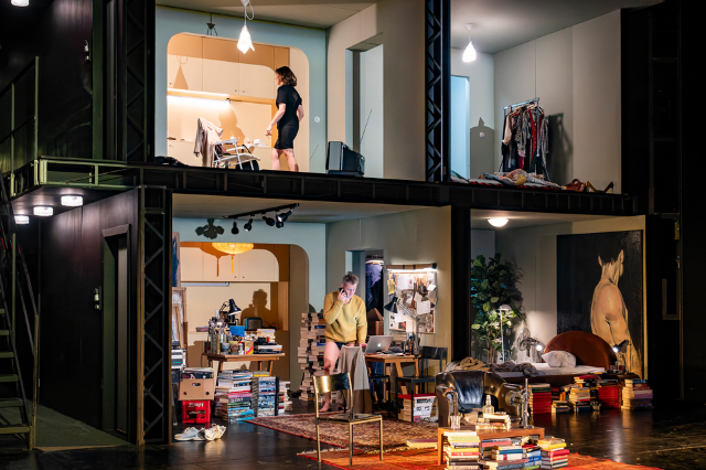 Ein zweistöckiges Bühnenbild zeigt Einblicke in mehrere Zimmer. Oben eine Frau im schlichten Raum, unten ein Mann zwischen Bücherstapeln am Laptop. Ein detaillierter Querschnitt durch private Lebenswelten.