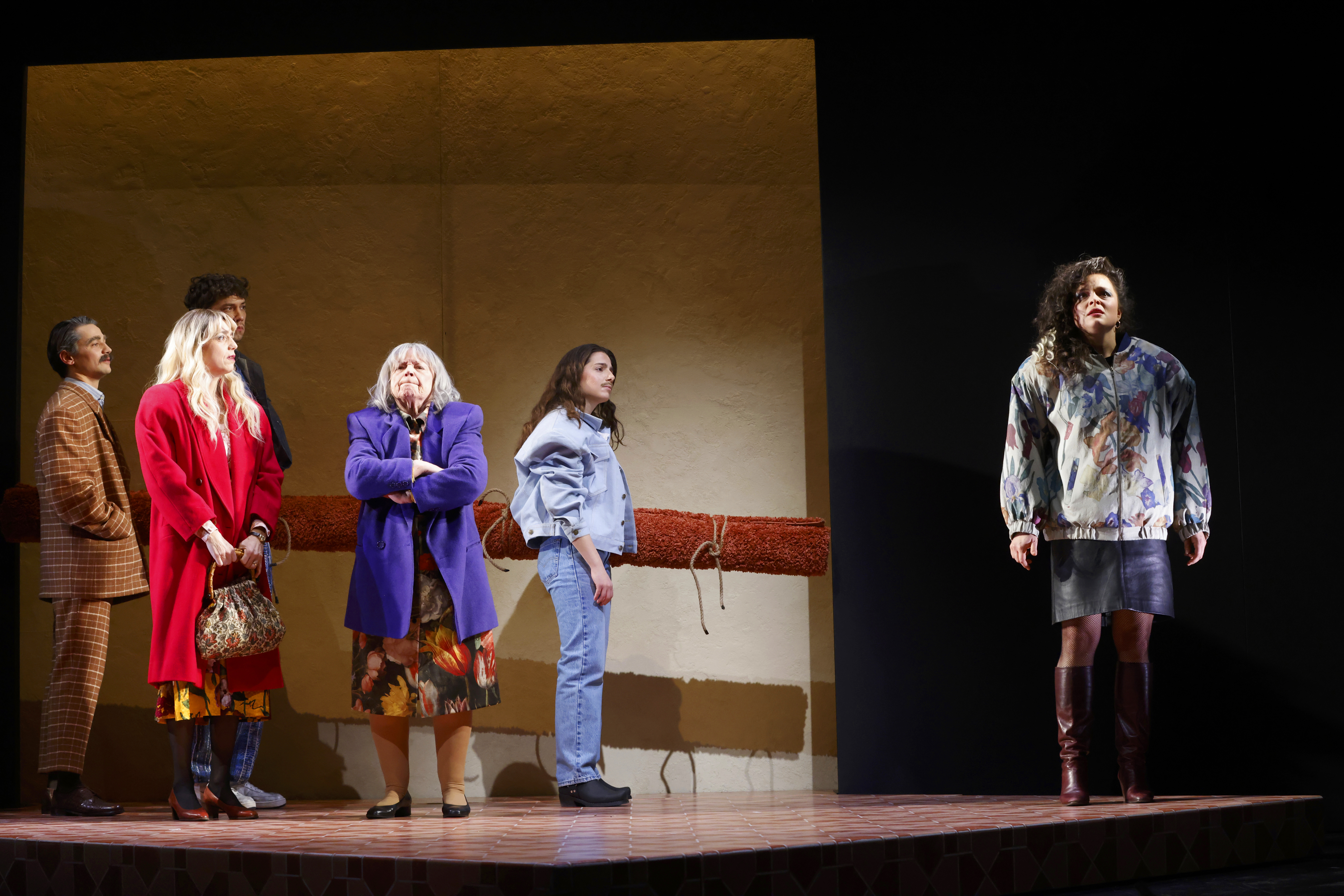 Fünf Personen auf einer Theaterbühne: Links stehen vier Personen in bunter Kleidung (u. a. roter Mantel, lila Sakko) vor einer hellen Wand. Rechts steht eine Frau isoliert im Rampenlicht und blickt auf.
