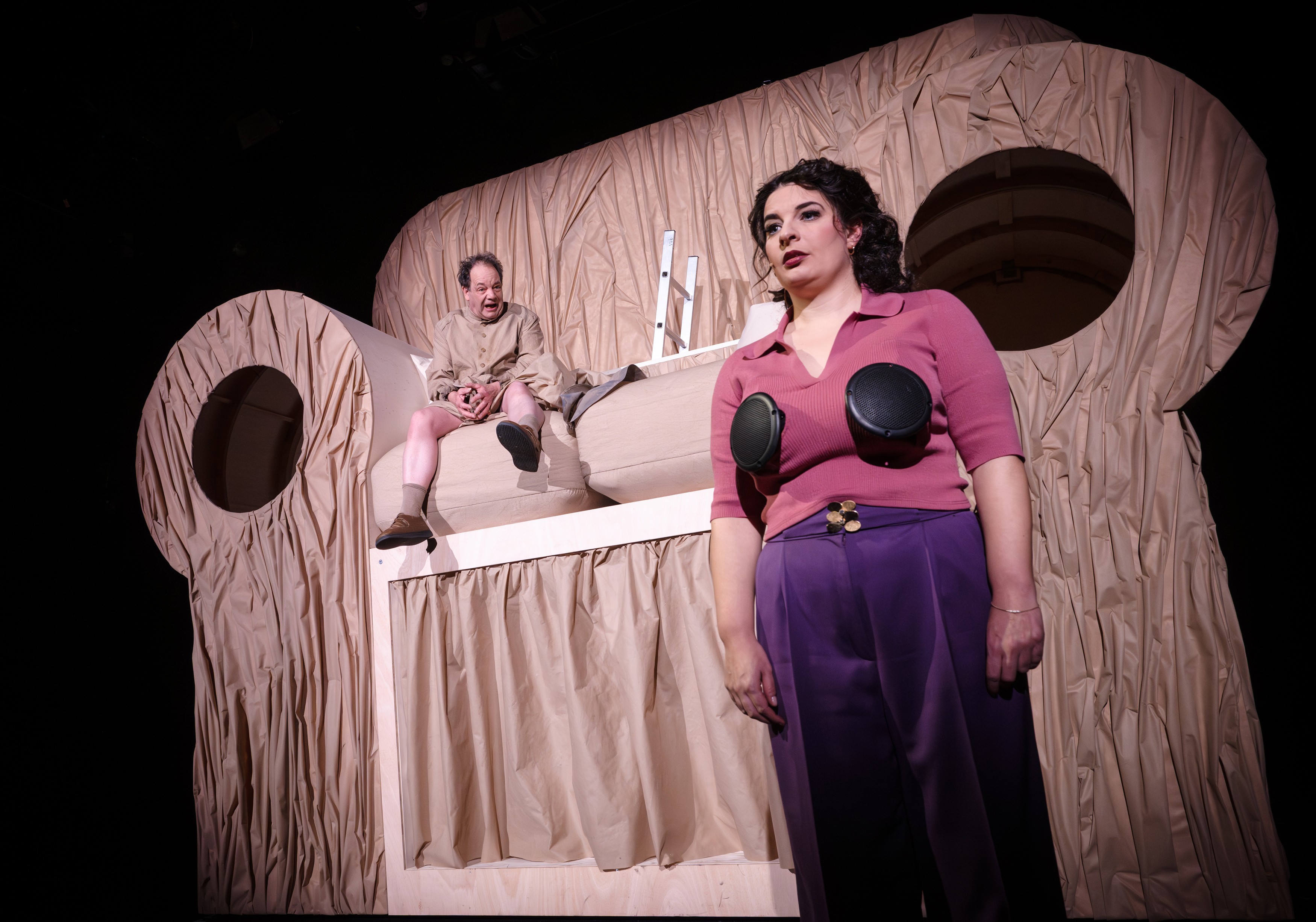 Zwei Personen auf einer Bühne vor einer riesigen, mit beigem Stoff drapierten Skulptur. Eine Frau im Vordergrund trägt Lautsprecher auf der Brust; ein Mann sitzt schreiend erhöht im Hintergrund.