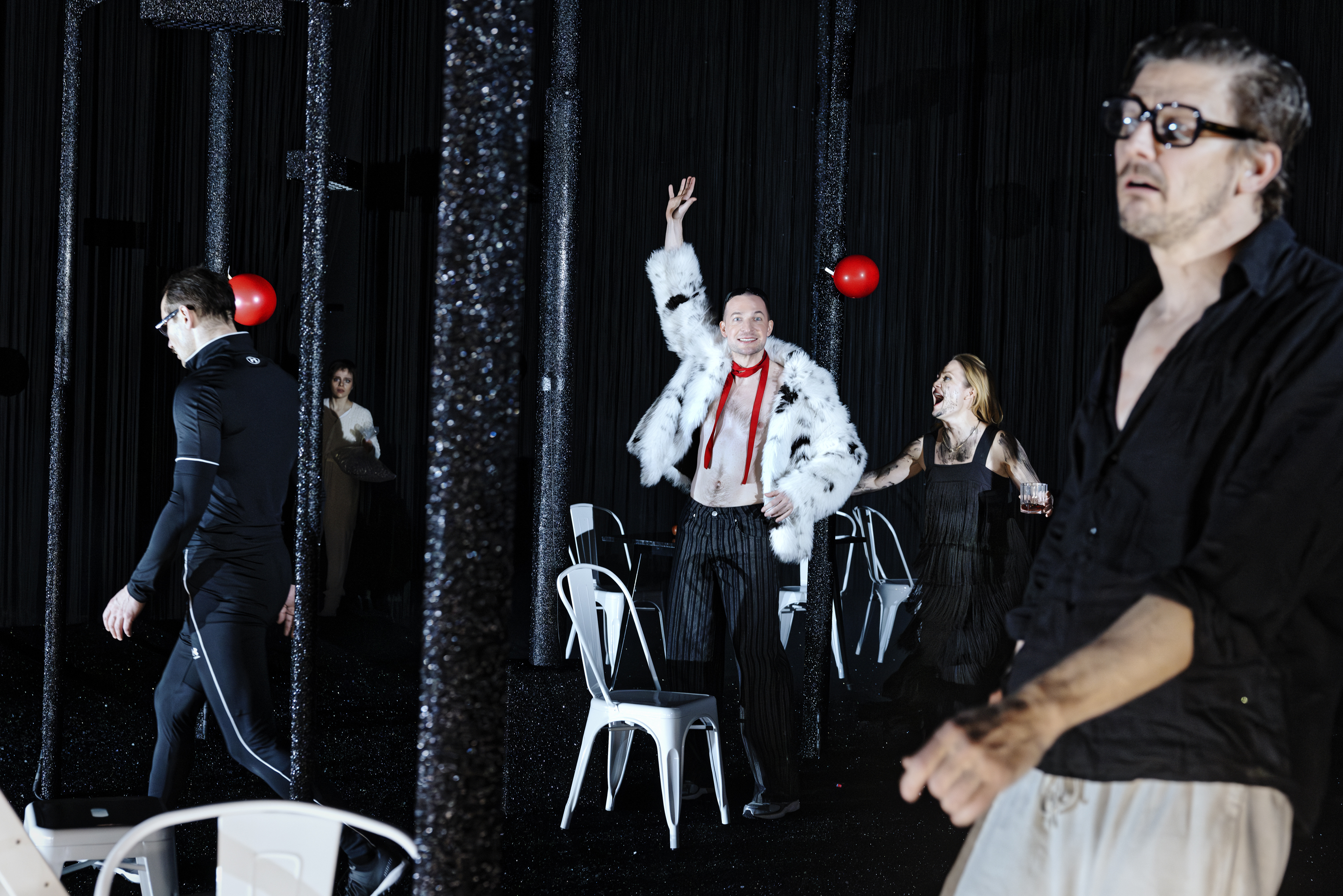 Düstere Bühnenszene aus „Kinder der Sonne“: Mehrere Personen agieren zwischen schwarzen Glitzersäulen. Mittig winkt ein Mann in Pelzjacke, rechts eine schreiende Frau, im Vordergrund ein Mann mit Brille.