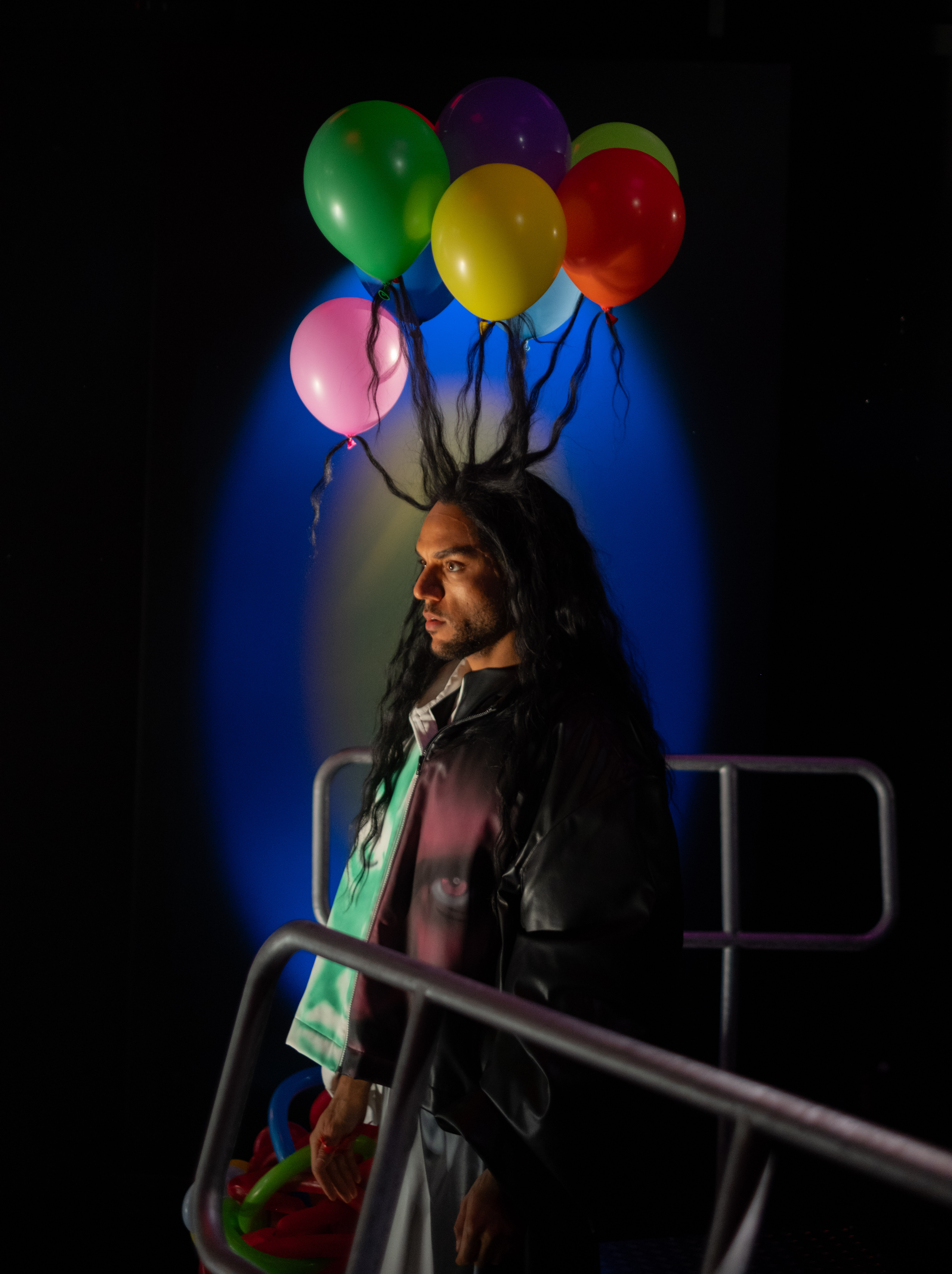 Mann mit langem Haar steht im Profil hinter einem Metallgeländer. Mehrere bunte Luftballons sind an seinen nach oben abstehenden Haarsträhnen befestigt. Im Hintergrund leuchtet ein blauer Lichtkegel.