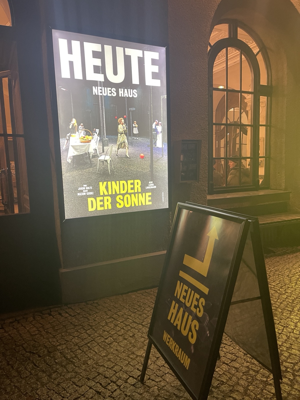 Ein beleuchteter Schaukasten zeigt das Theaterplakat „Kinder der Sonne“ im Neuen Haus. Davor steht ein Kundenstopper mit einem gelben Pfeil, der nach rechts oben Richtung „Neues Haus Werkraum“ weist.