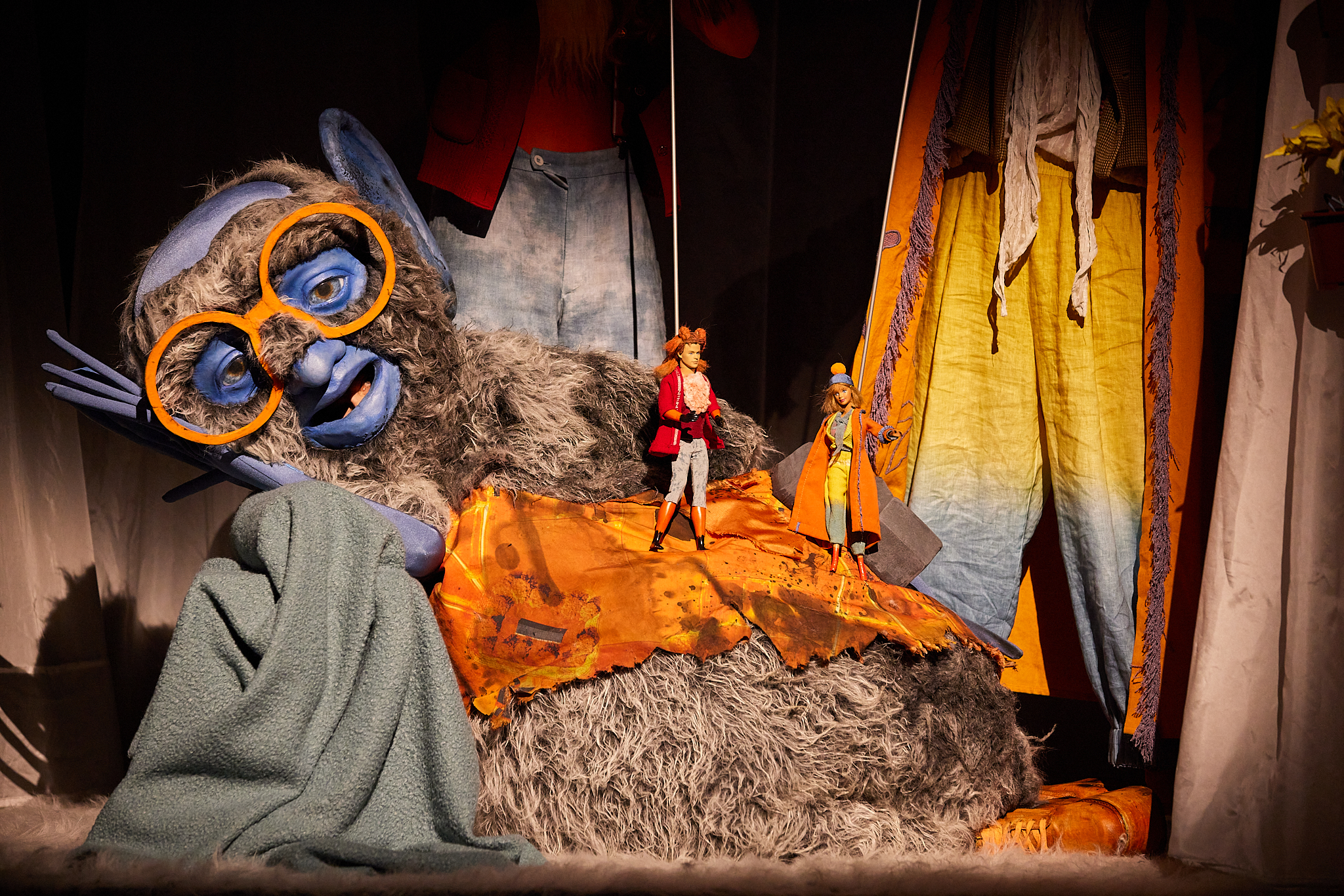 Theaterszene mit einer riesigen, grau-pelzigen Figur mit blauen Gesichtszügen und oranger Brille, die entspannt liegt. Auf ihrem Bauch stehen zwei kleine, modisch gekleidete Puppen vor buntem Hintergrund.
