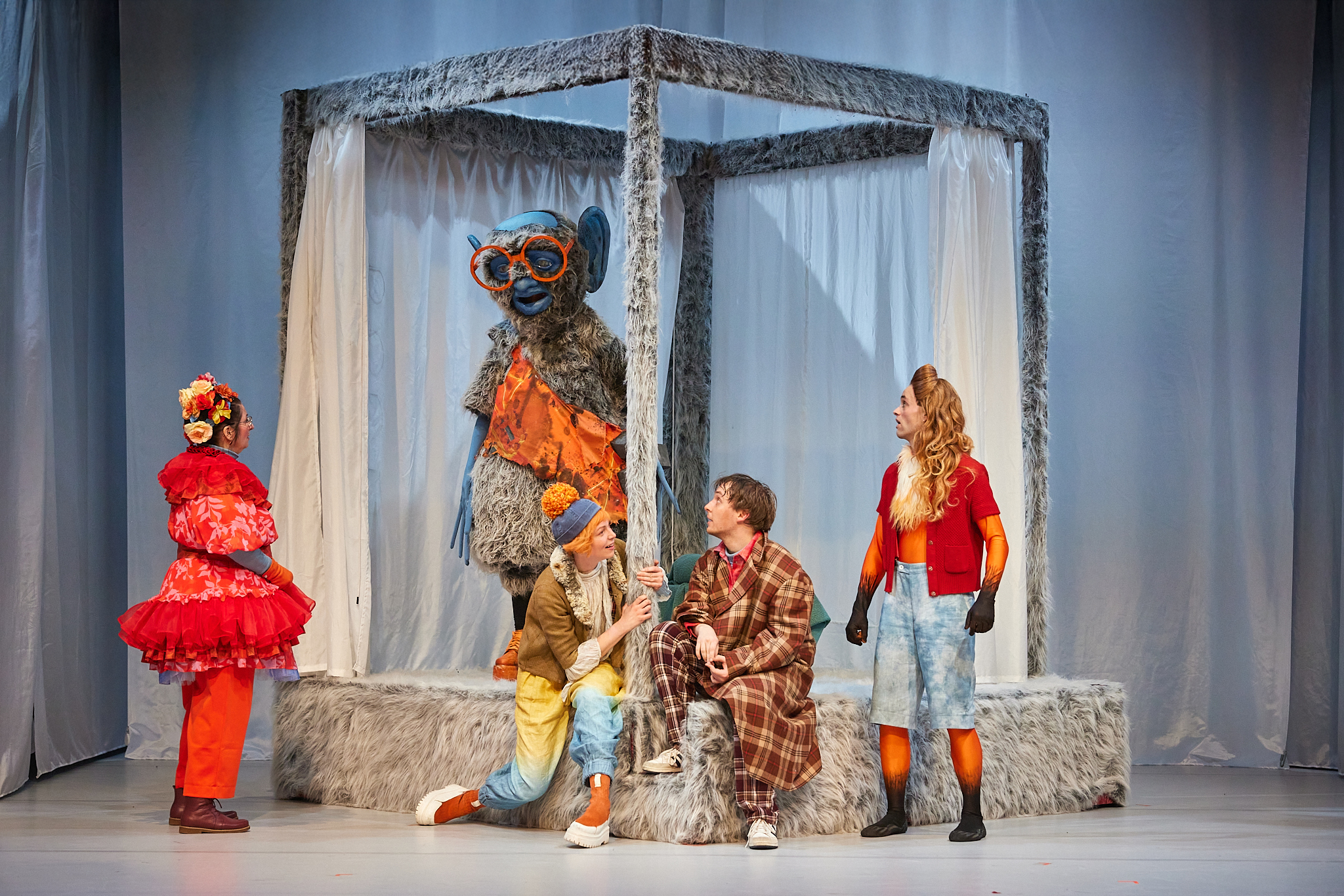 Theaterszene mit vier bunt kostümierten Schauspielern vor einem grauen, pelzigen Himmelbett-Gestell. Darin steht eine große, zottelige Figur mit blauem Gesicht und oranger Brille vor hellem Hintergrund.