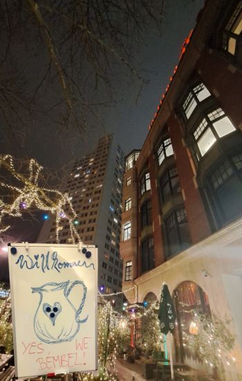 Abendliche Stadtszene: Ein Whiteboard mit der Aufschrift „Willkommen – YES, WE BEMBEL!“ und einer Krug-Zeichnung steht vor beleuchteten Bäumen und Häuserfassaden unter nächtlichem Himmel.