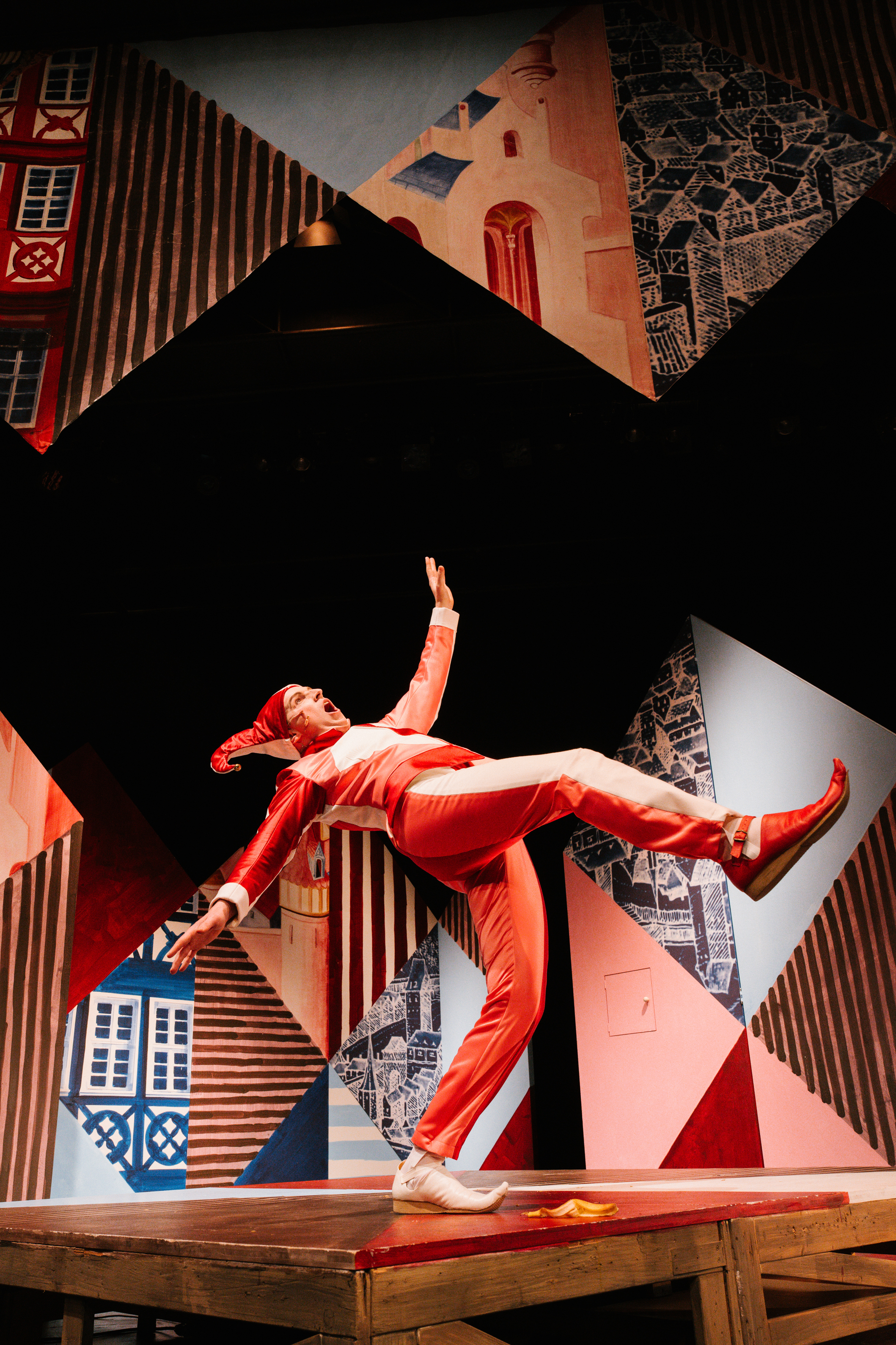 Bildbeschreibung Barrierefreiheit Benutzerdefiniertes Gem Bildbeschreibung Barrierefreiheit hat gesagt: Ein Schauspieler als Till Eulenspiegel in rot-weißem Kostüm und Narrenkappe balanciert dynamisch auf einem Bein auf einer Holzbühne. Im Hintergrund abstrakte, bunte Kulissenteile mit mittelalterlichen Häusern.