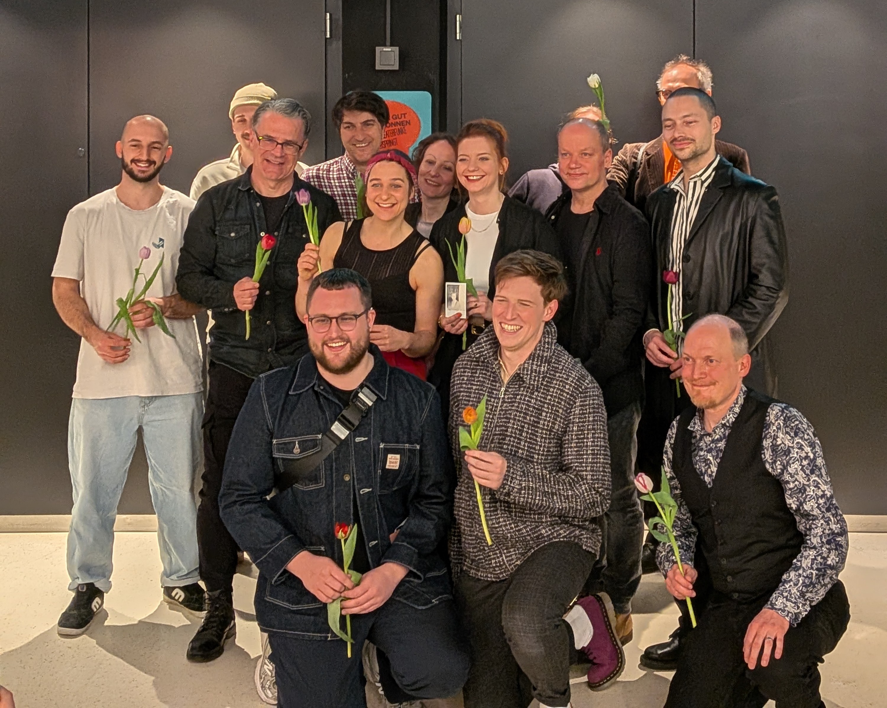 Eine Gruppe von zwölf lächelnden Erwachsenen posiert vor einer dunklen Wand. Viele halten bunte Tulpen in den Händen. Drei Personen knien im Vordergrund, die anderen stehen dicht gedrängt in zwei Reihen.