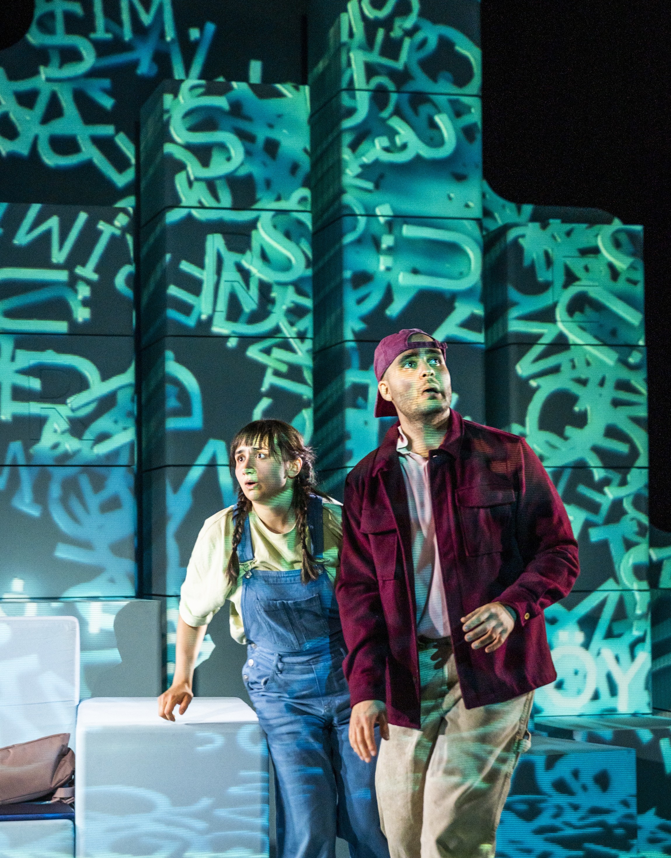Zwei Personen stehen auf einer Bühne vor hohen Quadern. Sie blicken gebannt nach oben. Ein Lichtprojektor wirft wirre, hellblaue Buchstaben über die gesamte Szene, die Personen und den Hintergrund.
