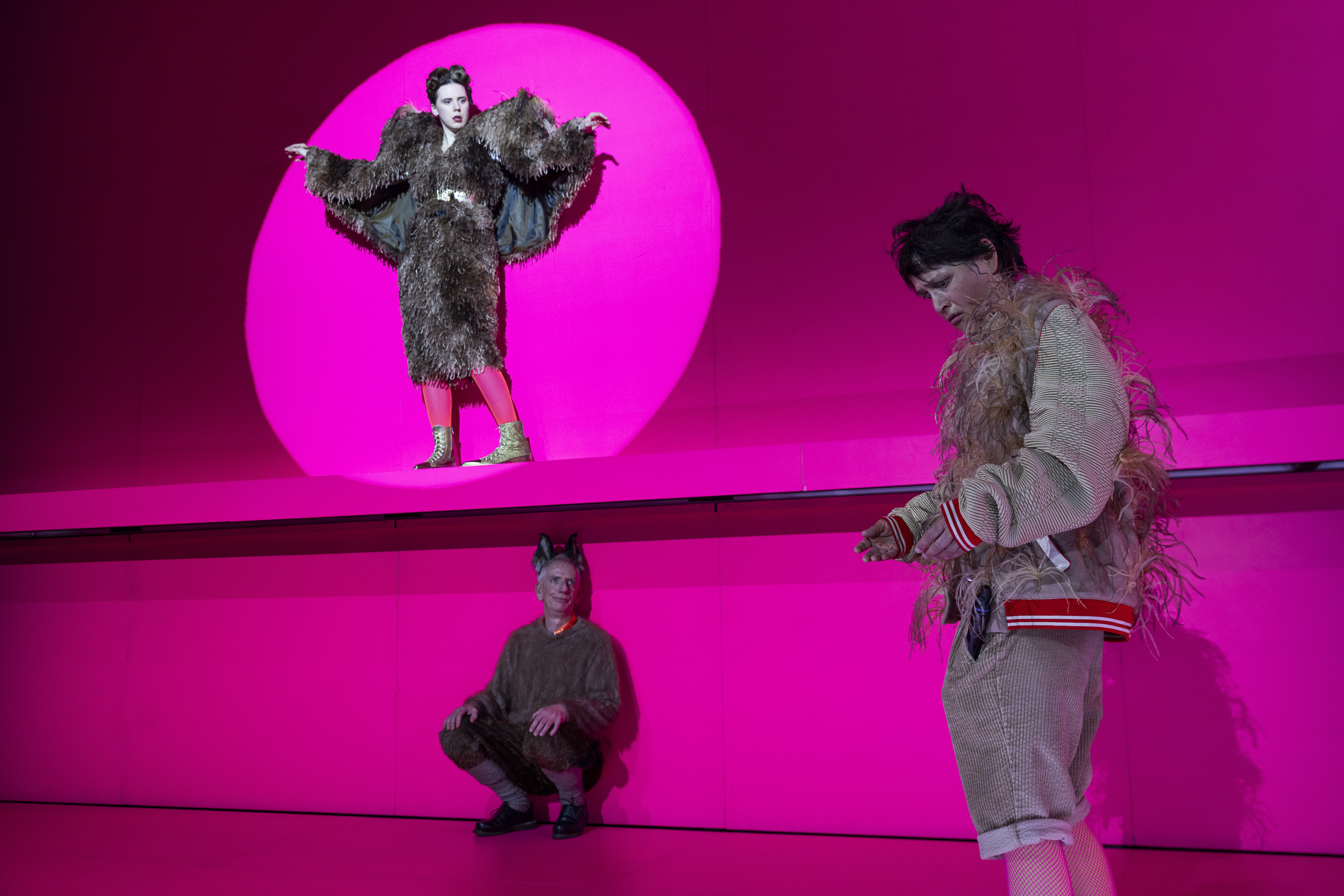Drei Personen auf einer pinken Bühne: Oben steht eine Performerin in einem braunen Federkostüm in einem Lichtkegel. Darunter kauert ein Mann mit Tierohren. Rechts blickt eine Person in Federjacke nach unten.