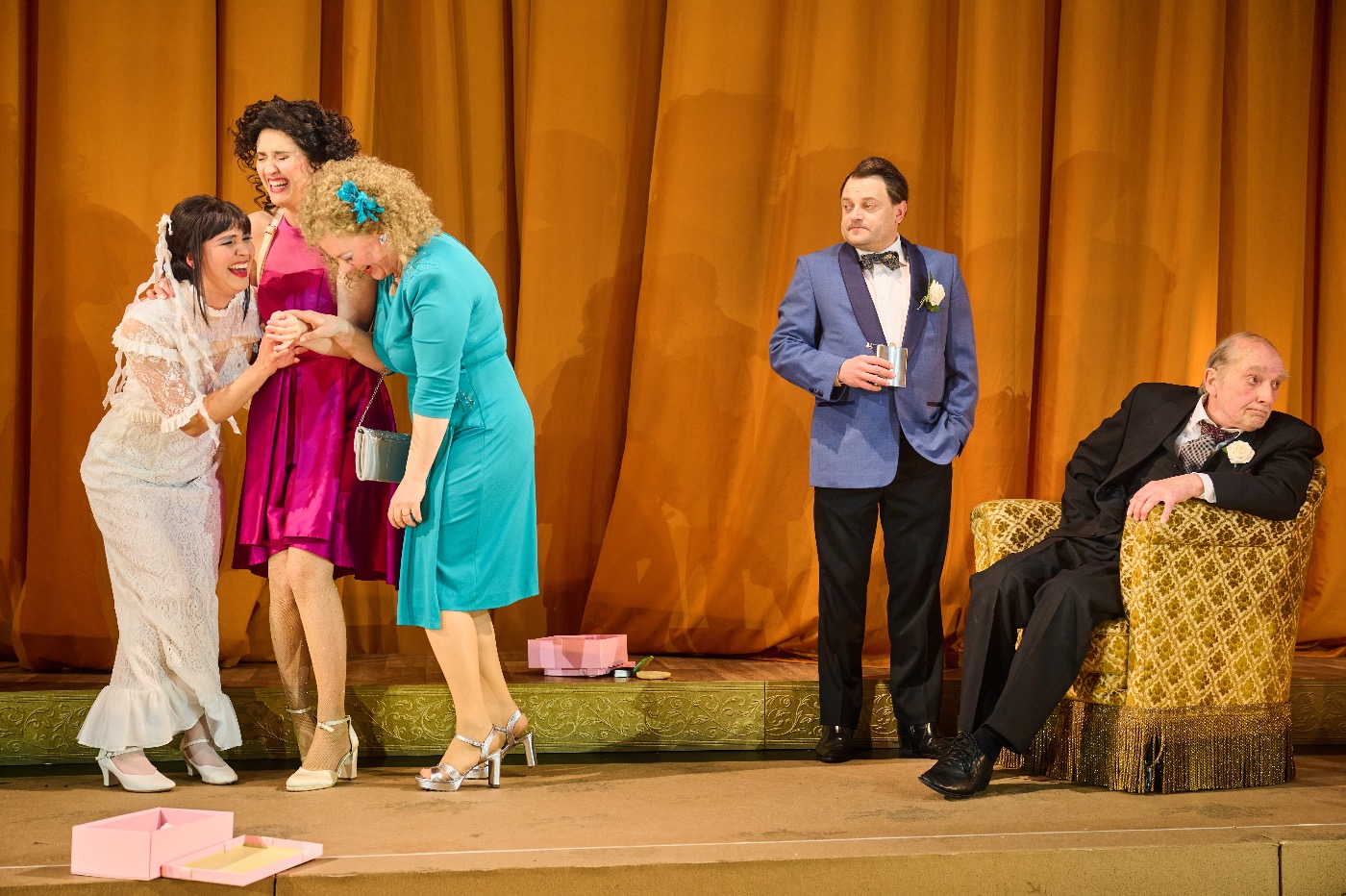 Drei lachende Frauen in festlichen Kleidern (weiß, pink, türkis) stehen links auf einer Bühne vor gelbem Vorhang. Rechts beobachtet ein Mann im blauen Sakko skeptisch einen älteren Herrn im Sessel.