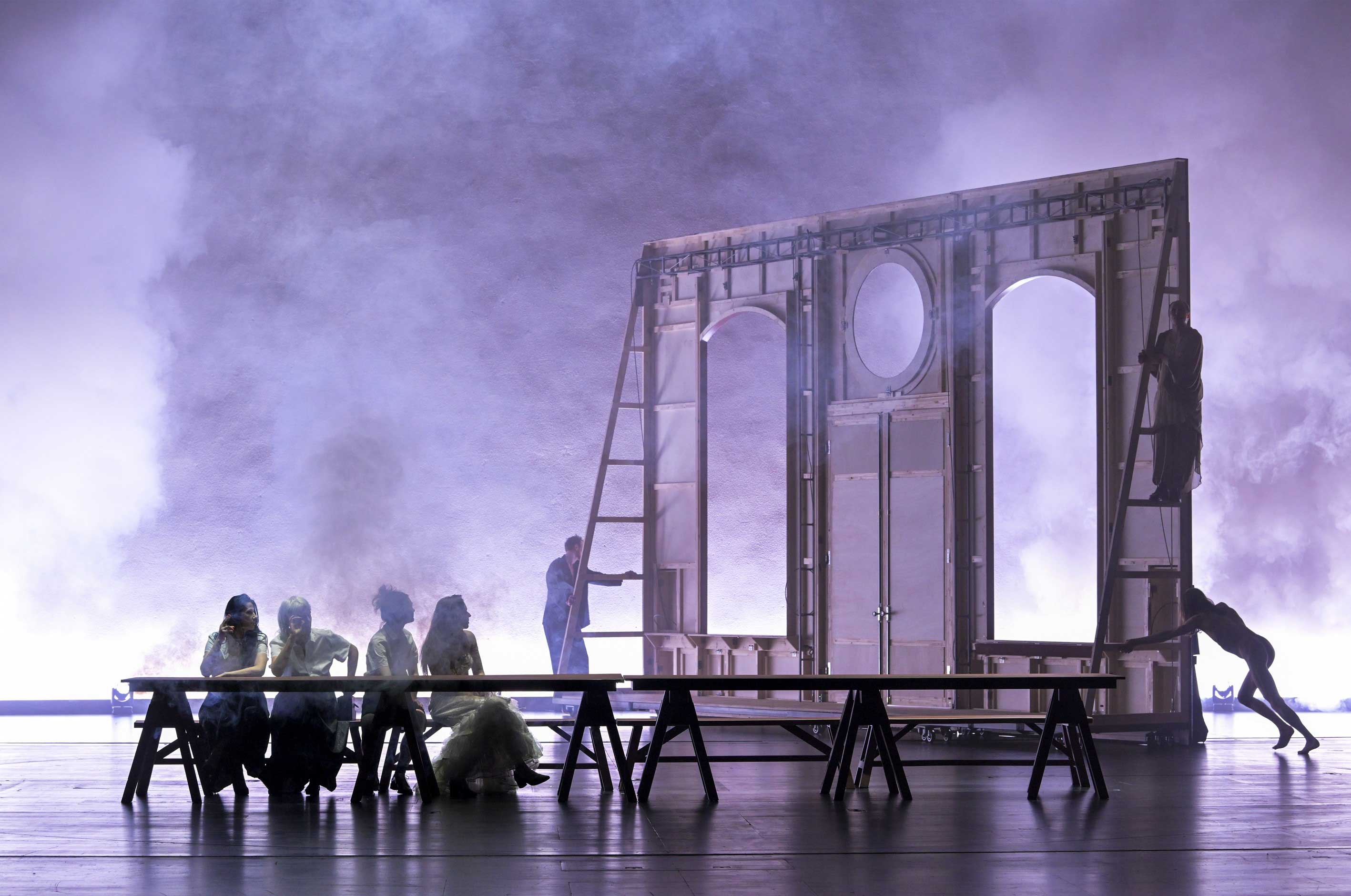 Eine Theaterbühne in violettem Licht und Nebel. Links sitzen vier Personen an einer langen Tafel. Rechts steht das Holzgerüst einer Fassade, an dem Personen lehnen und klettern. Mystische Atmosphäre.