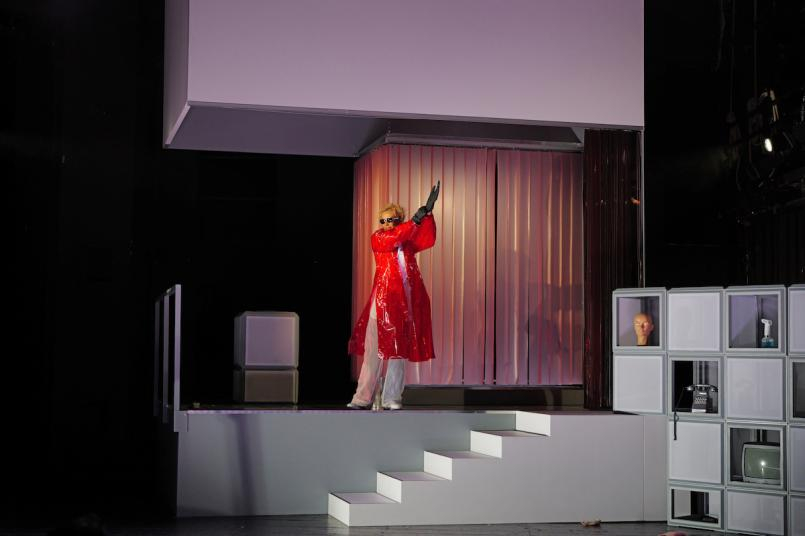 Theaterbühne mit minimalistischem Set: Eine Person im auffälligen roten Vinylmantel steht posierend auf einem weißen Podest mit Treppe vor einem hellrosa Vorhang. Rechts stehen quadratische weiße Regalboxen.