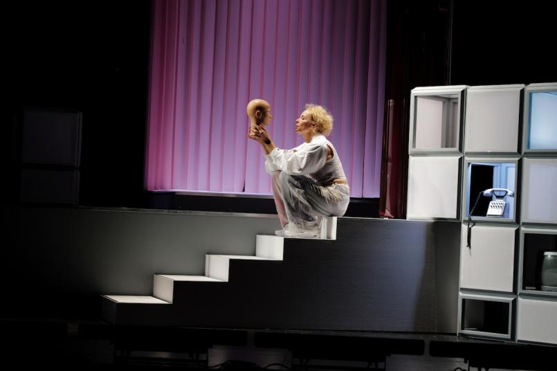 Eine blonde Person hockt auf einer weißen Treppe auf einer Theaterbühne und blickt auf einen Schaufensterpuppenkopf in ihrer Hand. Im Hintergrund ist ein rosa Vorhang, rechts ein modulares Regal mit Telefon.