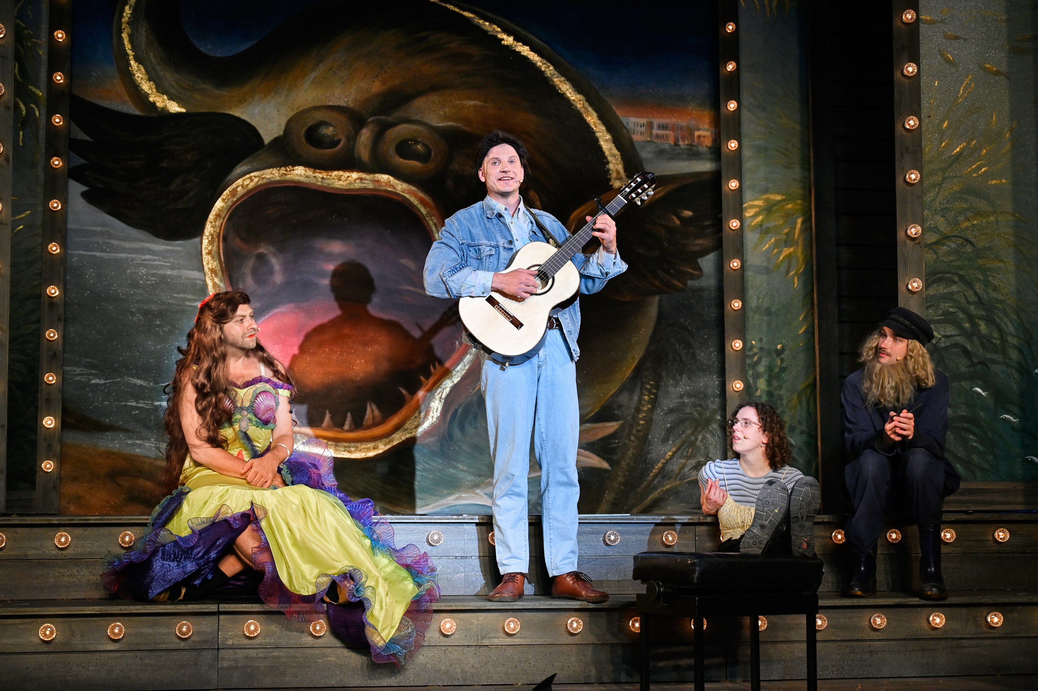 Theaterbühne: Ein Mann im Denim-Outfit singt und spielt Gitarre vor einem gemalten Walmaul. Links sitzt eine Person im bunten Kleid mit Perücke, rechts sitzen zwei weitere Schauspielende vor Kulissenlichtern.