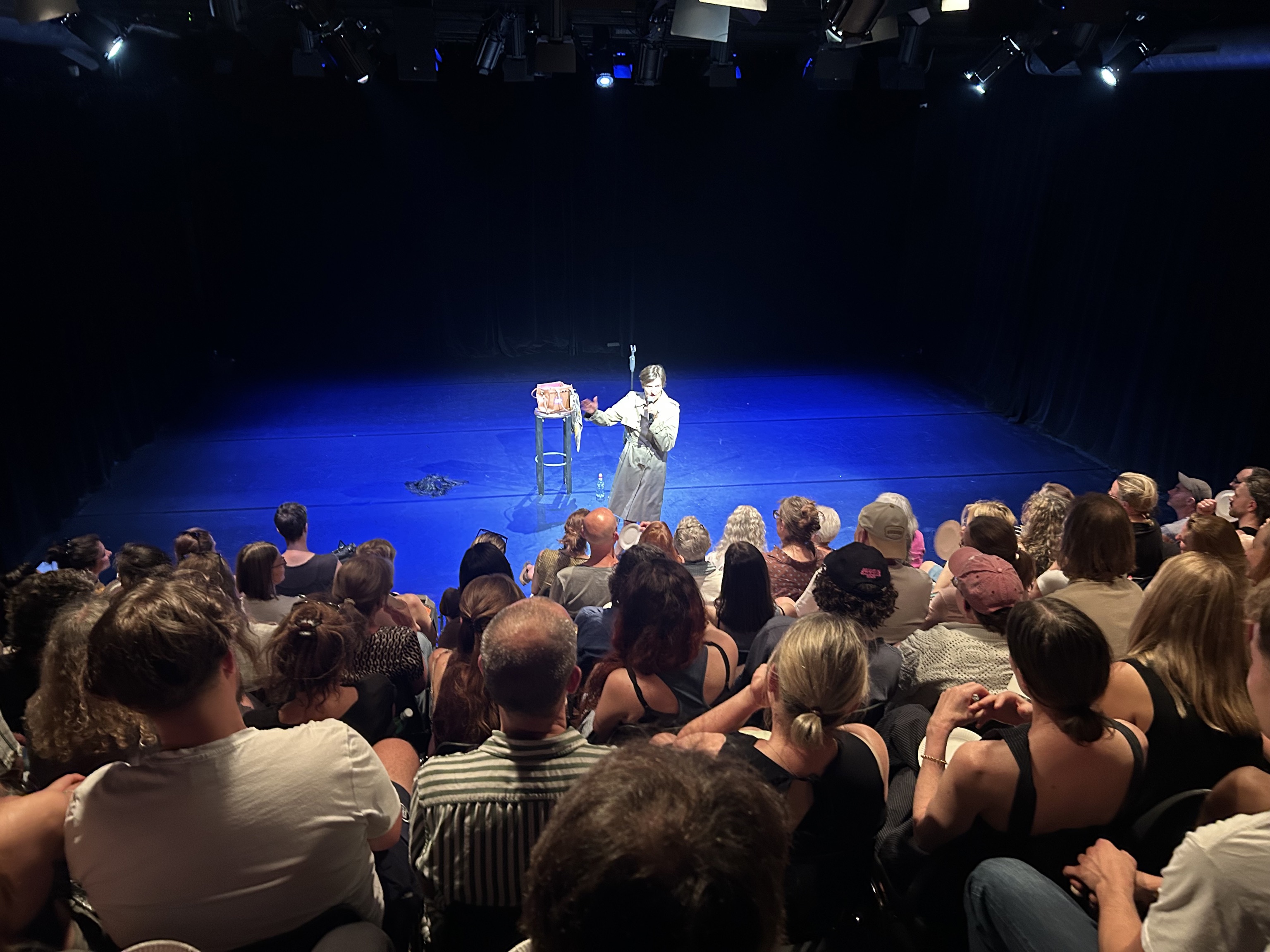 Blick von hinten auf ein Publikum im abgedunkelten Theatersaal, das auf eine intensiv blau beleuchtete Bühne schaut. Dort steht eine einzelne Person im hellen Trenchcoat neben einem Hocker im Scheinwerferlicht.