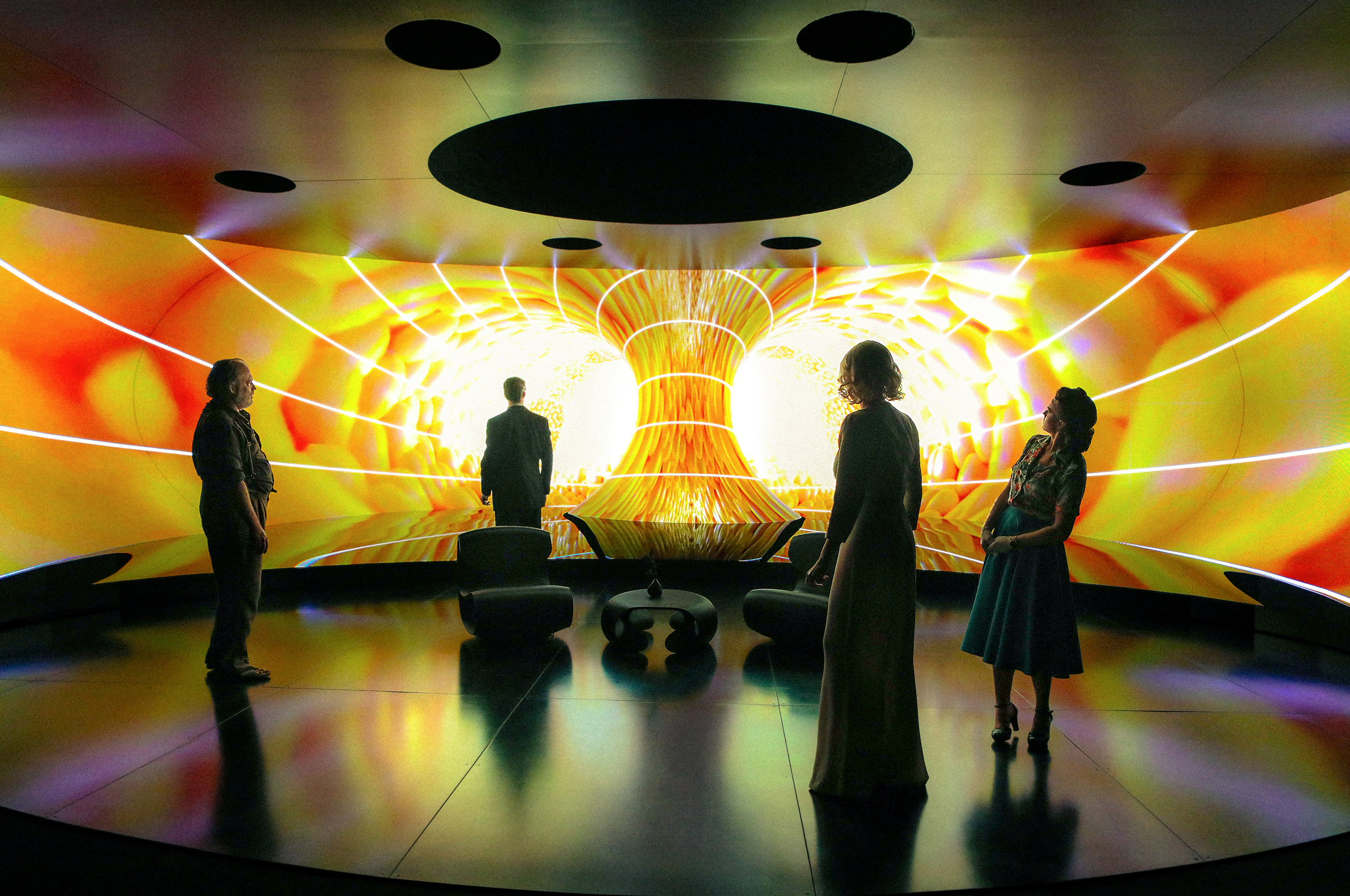 Vier Personen stehen in einem abgedunkelten, runden Raum mit spiegelndem Boden. Sie betrachten eine riesige Panoramaleinwand, die ein grell leuchtendes, orange-gelbes Energiefeld in Form eines Tunnels zeigt.