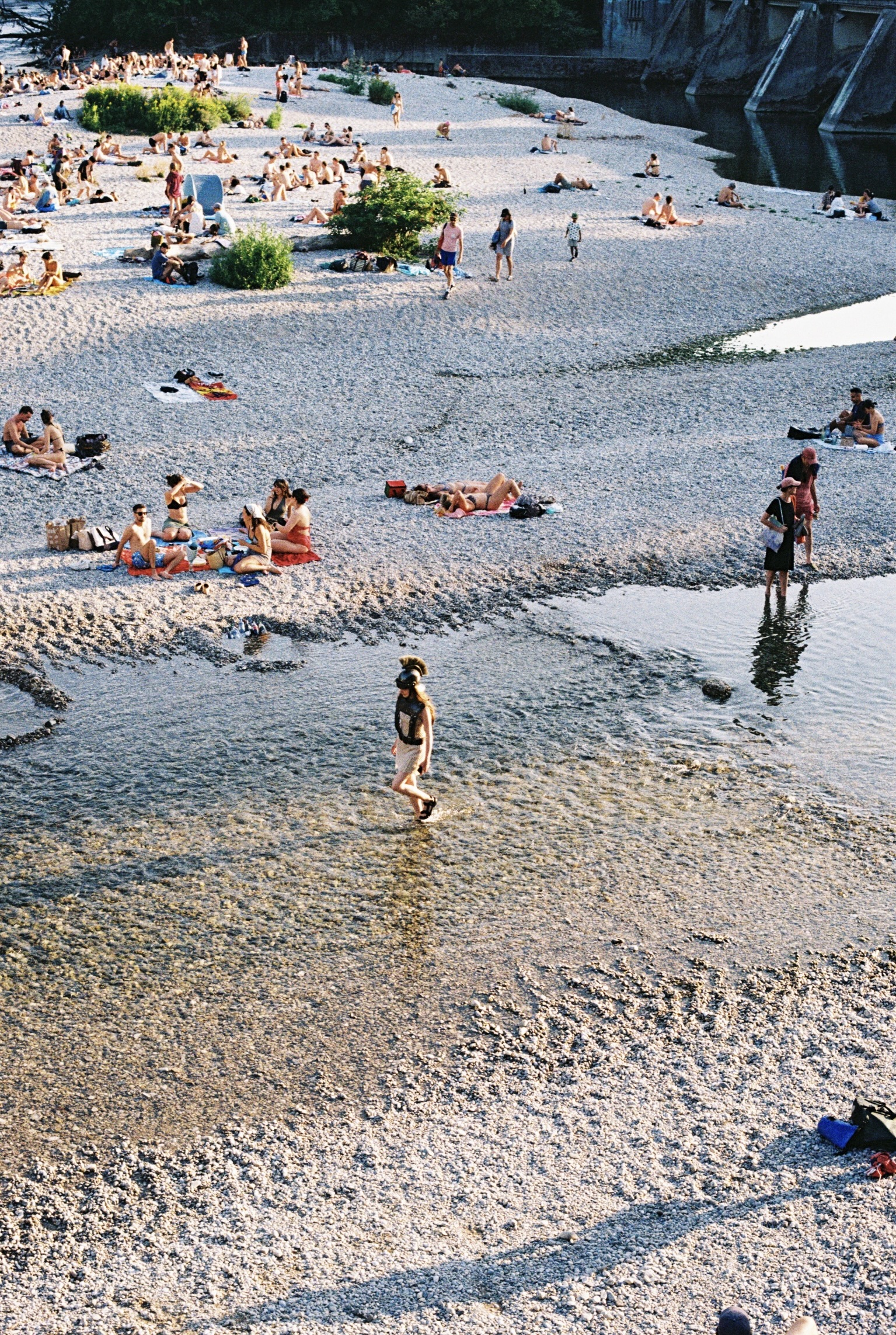 Eine Frau in griechischer Rüstung mit Helm watet durch das flache Wasser an einem belebten Kieselstrand. Im Hintergrund entspannen sich viele Badegäste an einem sonnigen Sommertag auf einem steinigen Flussufer.