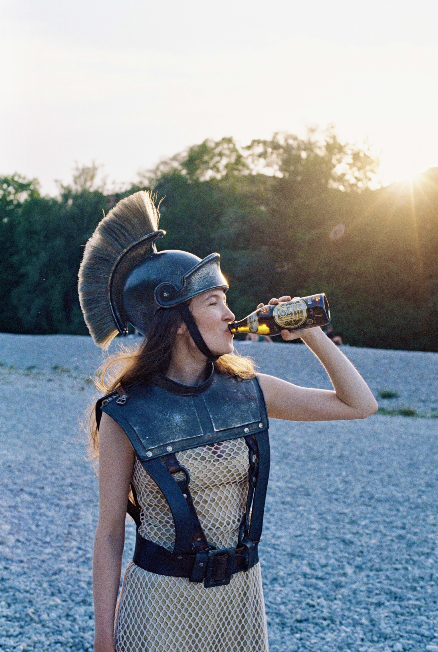 Eine junge Frau in griechischer Rüstung, bestehend aus Brustpanzer und Helm mit Helmbusch, steht an einem Kieselstrand. Im Gegenlicht der goldenen Abendsonne trinkt sie genüsslich aus einer braunen Bierflasche.