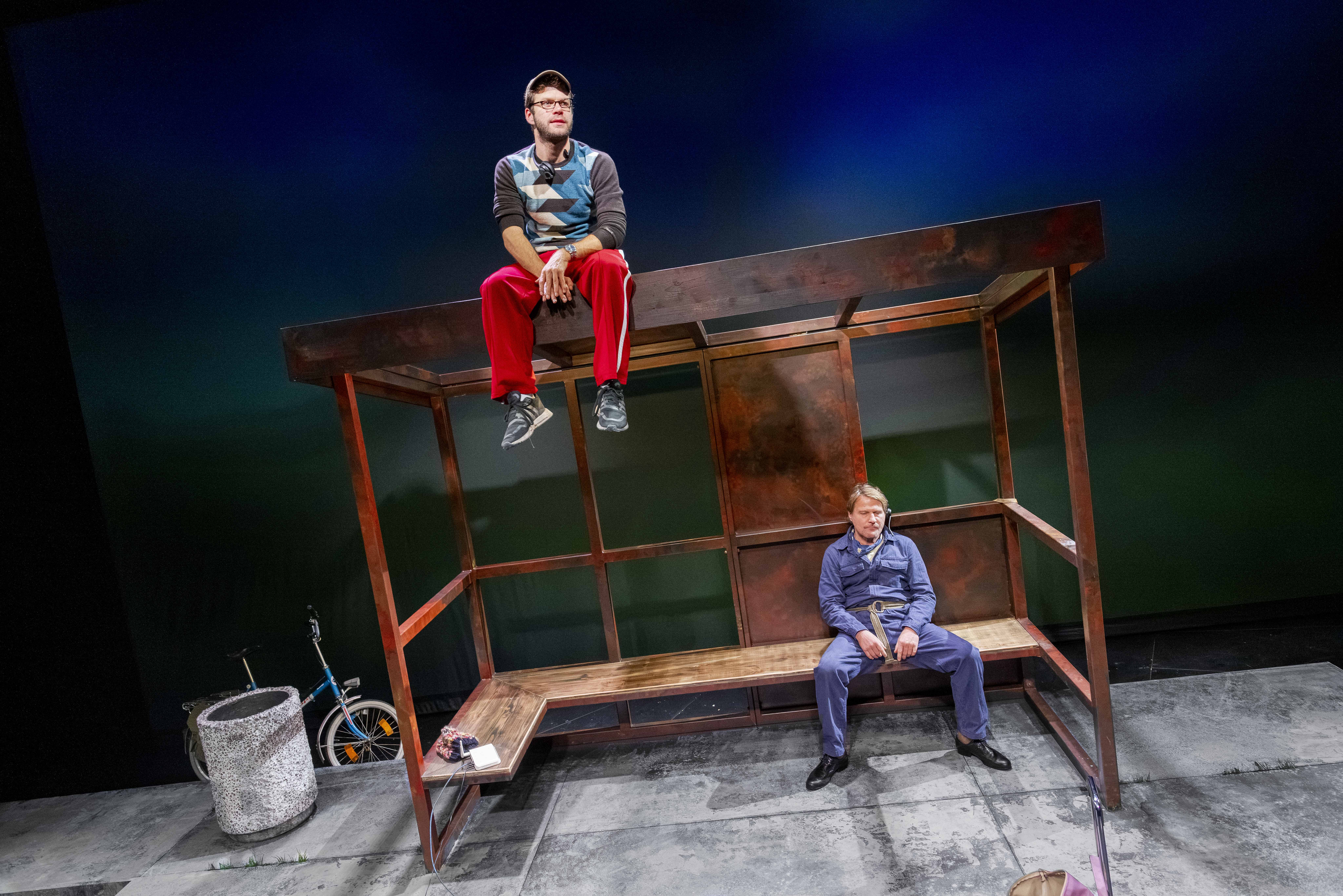 Auf einer Theaterbühne steht eine rostige Bushaltestelle. Ein Mann in roter Jogginghose und Kappe sitzt oben auf dem Dach, während ein zweiter im blauen Overall gelassen unten auf der hölzernen Bank sitzt.